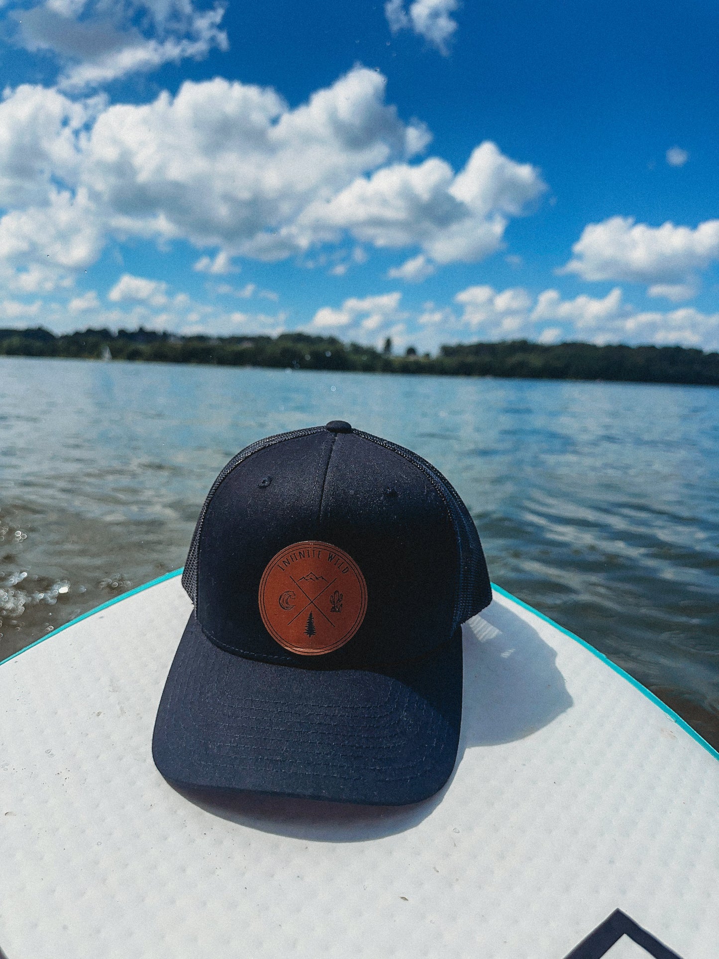 Brown trucker snap back with brown leather infinite wild patch on the front. The hat sits on top of a paddle boarding in the middle of a lake on a partially sunny day with blue skies and big puffy clouds